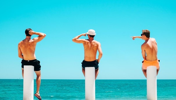 Men wearing swimwear, sitting at the beach looking out to sea.